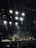 A concert scene with a band performing on stage under a series of bright spotlights. Smoke effects enhance the atmosphere as the audience watches. Musical instruments like guitars, keyboards, and drums are visible, and the performers are silhouetted against the illuminated background.
