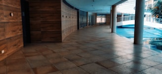 A modern indoor hallway with tiled flooring and wooden panel walls. The space features large windows allowing natural light to enter, creating reflections on the floor. A curved ceiling and multiple pillars add to the architectural interest. A bridge-like structure is visible in the background alongside greenery.