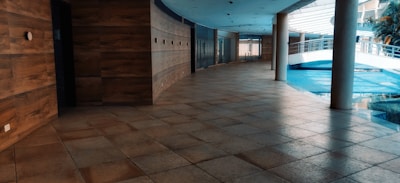 A modern indoor hallway with tiled flooring and wooden panel walls. The space features large windows allowing natural light to enter, creating reflections on the floor. A curved ceiling and multiple pillars add to the architectural interest. A bridge-like structure is visible in the background alongside greenery.