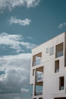 A modern, minimalist concrete building stands against a backdrop of a clear blue sky with scattered fluffy white clouds. The structure features large, angular windows and open balconies with glass railings, providing a sense of openness and simplicity.