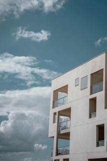 A modern, minimalist concrete building stands against a backdrop of a clear blue sky with scattered fluffy white clouds. The structure features large, angular windows and open balconies with glass railings, providing a sense of openness and simplicity.