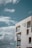 A modern, minimalist concrete building stands against a backdrop of a clear blue sky with scattered fluffy white clouds. The structure features large, angular windows and open balconies with glass railings, providing a sense of openness and simplicity.