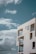 A modern, minimalist concrete building stands against a backdrop of a clear blue sky with scattered fluffy white clouds. The structure features large, angular windows and open balconies with glass railings, providing a sense of openness and simplicity.