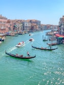 A picturesque canal bordered by historic Venetian buildings on either side, with several gondolas carrying passengers in the turquoise water. Brightly colored awnings and bustling activity suggest a lively atmosphere.