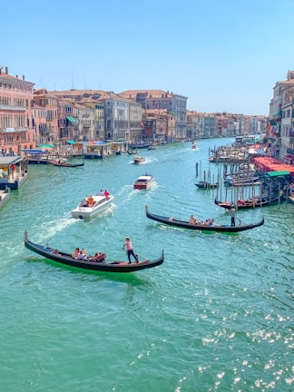 A picturesque canal bordered by historic Venetian buildings on either side, with several gondolas carrying passengers in the turquoise water. Brightly colored awnings and bustling activity suggest a lively atmosphere.