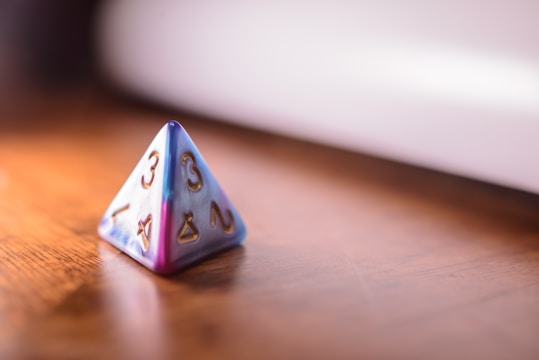 A four-sided die (tetrahedron) with numbers and symbols on each face, placed on a wooden surface. The die has a visually striking color gradient that transitions from blue to purple, and the numbers and symbols are engraved and highlighted in gold.