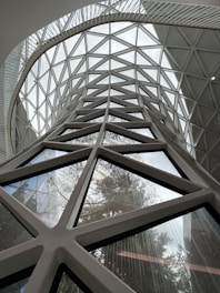 A modern architectural structure featuring large triangular glass panels forming a lattice design. The structure allows an abundance of natural light and views of trees outside, visible through the glass. The geometric pattern creates a dynamic and complex visual experience.
