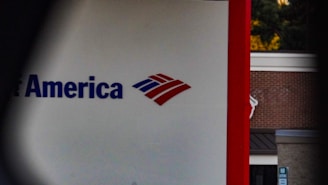 A partial view of a sign, displaying a recognizable red, white, and blue logo. The words 'of America' are visible, suggesting it is associated with a financial institution. The background shows a building with a brick facade.