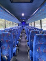 The interior of a bus is depicted, with two rows of blue, patterned seats lined up along a central aisle. The windows on either side allow natural light to illuminate the vibrant upholstery.
