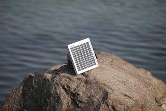 A solar panel is positioned on a large rock near a body of water, suggesting energy harnessing in a natural setting.