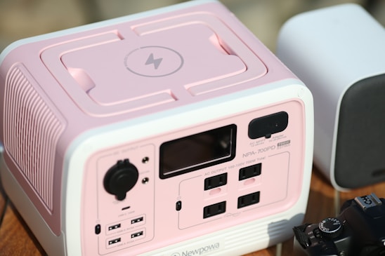 A pink and white power station with multiple ports and a black knob on the front panel. The device has a display and is labeled as NPA-700PD with 700W power capacity. In the background, there is a white object with rounded edges, possibly a speaker, and a black camera partially visible at the bottom right.