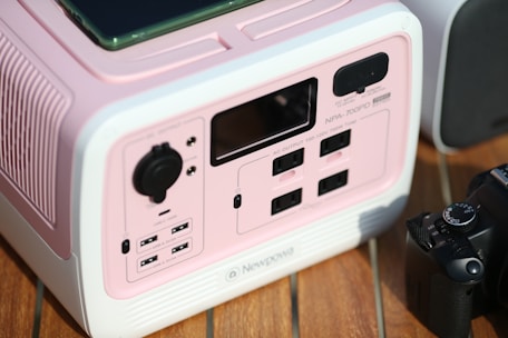 A pink and white electronic device featuring multiple power outlets, USB ports, and control knobs is placed on a wooden surface. A black camera is positioned next to it.
