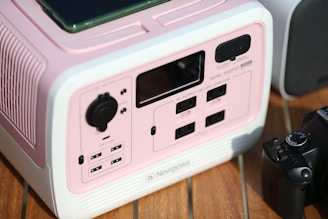 A pink and white electronic device featuring multiple power outlets, USB ports, and control knobs is placed on a wooden surface. A black camera is positioned next to it.