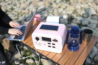 A portable power station is placed on a wooden table, with a smartphone charging on top of it. Nearby, there is a blue lantern and a metal cup. A person is working on a laptop next to the power station, with a stone surface in the background.