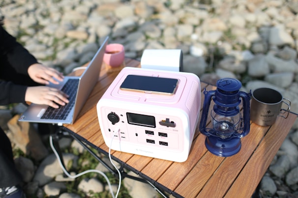 A portable power station is placed on a wooden table, with a smartphone charging on top of it. Nearby, there is a blue lantern and a metal cup. A person is working on a laptop next to the power station, with a stone surface in the background.