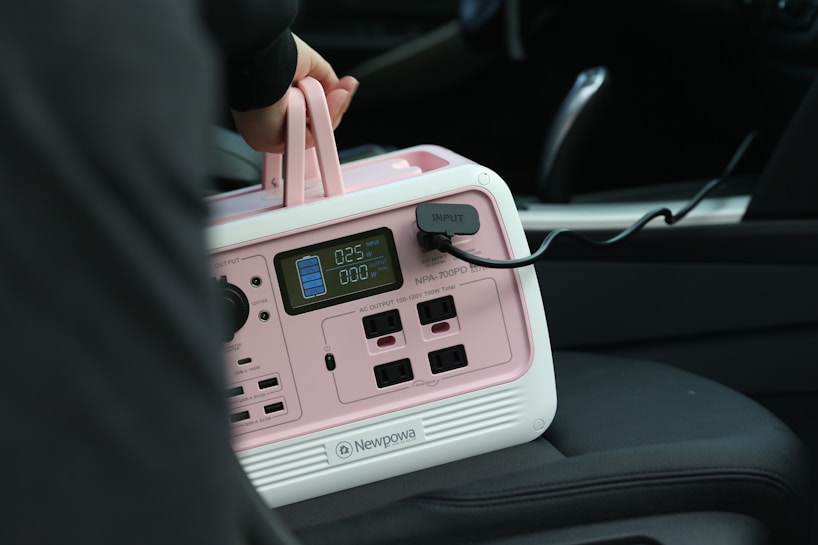 A portable power station with a pink and white casing is placed on a car seat. A person's hand is holding its handle. The device has multiple outlets and a digital display showing power metrics. A cable is plugged into the input socket.