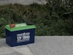 A blue 12V 100Ah lithium battery with green top, featuring red and black cable clamps, is placed on a concrete surface. Surrounding the battery are lush green shrubs and plants.