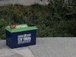 A blue 12V 100Ah lithium battery with green top, featuring red and black cable clamps, is placed on a concrete surface. Surrounding the battery are lush green shrubs and plants.