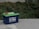 A blue 12V 100Ah lithium battery with green top, featuring red and black cable clamps, is placed on a concrete surface. Surrounding the battery are lush green shrubs and plants.