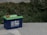 A blue 12V 100Ah lithium battery with green top, featuring red and black cable clamps, is placed on a concrete surface. Surrounding the battery are lush green shrubs and plants.