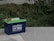 A blue 12V 100Ah lithium battery with green top, featuring red and black cable clamps, is placed on a concrete surface. Surrounding the battery are lush green shrubs and plants.