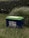 A lithium battery labeled 12V 100Ah is situated on grass, with its green top contrasting with its dark blue body. The background features a thick hedge, adding a natural setting to the image.