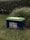 A lithium battery labeled 12V 100Ah is situated on grass, with its green top contrasting with its dark blue body. The background features a thick hedge, adding a natural setting to the image.