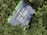 A 12V 100Ah lithium battery with the branding Newpowa is surrounded by dense green foliage. The battery case is blue, and it prominently displays technical specifications, along with website and contact details.