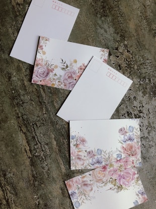Several postcards are arranged on a textured surface. Each postcard features floral designs with pastel-colored flowers, including pinks, purples, and blues. Some postcards have a designated area for address and stamps, outlined in red.