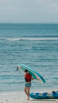 A person wearing a red and black life vest stands on a sandy beach near the ocean. They carry a turquoise and white paddleboard on their shoulder. The sea is calm with gentle waves, and there is another inflatable object in the foreground resting on the sand.