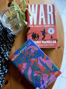 A wooden table holds two books and a jar with a plant. The book titled 'WAR: How Conflict Shaped Us' has a bold red cover with silhouettes of soldiers, and 'Grief is Love: Living with Loss' features a vibrant, abstract design with blues and pinks. A patterned fabric is partially visible underneath the jar.