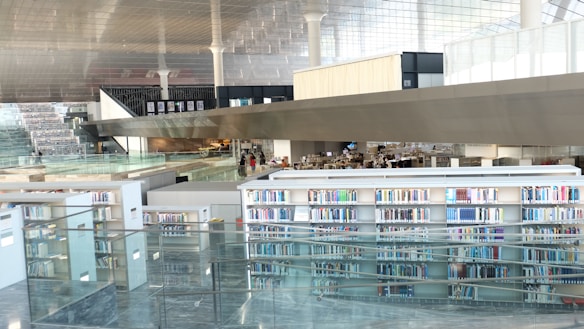A modern library interior with large glass windows and multiple levels of bookshelves filled with colorful books. The design is sleek and open, featuring a variety of study areas and contemporary architectural elements like glass railings and a reflective ceiling.