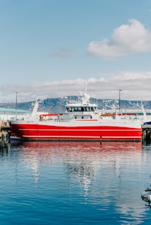 A large red and white fishing vessel docked at a harbor, with clear blue water reflecting its image. The background features snowy mountains under a partly cloudy sky, creating a serene and picturesque setting.