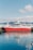 A large red and white fishing vessel docked at a harbor, with clear blue water reflecting its image. The background features snowy mountains under a partly cloudy sky, creating a serene and picturesque setting.