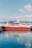 A large red and white fishing vessel docked at a harbor, with clear blue water reflecting its image. The background features snowy mountains under a partly cloudy sky, creating a serene and picturesque setting.