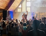 A lively indoor music concert with a singer on stage, engaging an enthusiastic crowd. The audience has their hands raised in excitement and participation. The setting is a wooden interior with bright stage lights and a screen displaying lyrics.