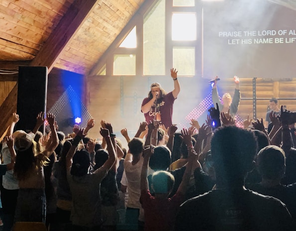 A lively indoor music concert with a singer on stage, engaging an enthusiastic crowd. The audience has their hands raised in excitement and participation. The setting is a wooden interior with bright stage lights and a screen displaying lyrics.