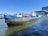 A large cargo boat named 'Wabo 3' floats on a waterway. The front is painted blue, while the rest of the boat shows signs of rust and age. In the background, there's a modern glass and steel building, and a large cruise ship is partially visible. The sky is clear and blue, and a person is standing on the deck of the boat.