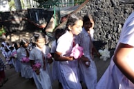 A group of young students in uniform are walking up a flight of stairs. They are wearing white dresses with red and white striped ties, carrying flowers in their hands. The background features a stone wall and sunlight casting shadows on the steps.