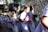 A group of young students in uniform are walking up a flight of stairs. They are wearing white dresses with red and white striped ties, carrying flowers in their hands. The background features a stone wall and sunlight casting shadows on the steps.
