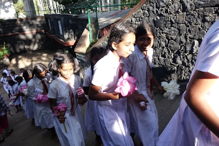 A group of young students in uniform are walking up a flight of stairs. They are wearing white dresses with red and white striped ties, carrying flowers in their hands. The background features a stone wall and sunlight casting shadows on the steps.
