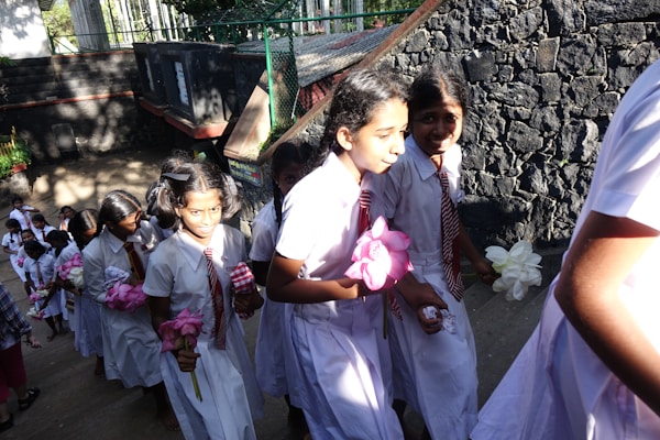 A group of young students in uniform are walking up a flight of stairs. They are wearing white dresses with red and white striped ties, carrying flowers in their hands. The background features a stone wall and sunlight casting shadows on the steps.
