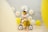 A toddler wearing a crown and a yellow dress is riding a small bike indoors. The decoration includes yellow and white balloons, with some shaped like daisies, and a large number '2' balloon in the background. The walls have some scribbles, possibly drawn by a child.
