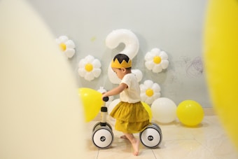 A toddler wearing a crown and a yellow dress is riding a small bike indoors. The decoration includes yellow and white balloons, with some shaped like daisies, and a large number '2' balloon in the background. The walls have some scribbles, possibly drawn by a child.