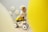 A young child wearing a yellow skirt and a party hat is sitting on a small white and black ride-on toy. The background features balloon decorations designed to look like daisies, and two large yellow balloons are in the foreground, partially obscuring the view.