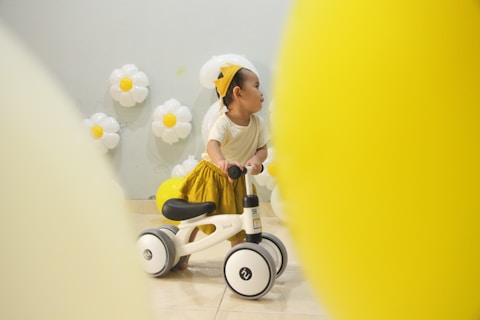 A young child wearing a yellow skirt and a party hat is sitting on a small white and black ride-on toy. The background features balloon decorations designed to look like daisies, and two large yellow balloons are in the foreground, partially obscuring the view.