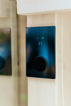 A modern, sleek black thermostat mounted on a light wooden wall. The digital display shows the number 21 prominently.