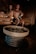 A dimly lit sauna room features a man pouring water over hot stones with a metal ladle, creating steam. A woman sits nearby, both dressed in swimwear, enjoying the relaxing atmosphere. The interior has wooden walls and benches, contributing to the warm and cozy environment.