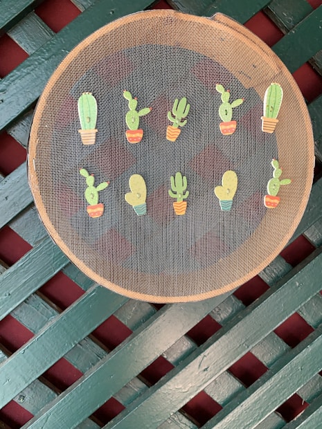 A circular mesh screen is decorated with small colorful illustrations of potted cacti. The screen is set against a background featuring a crisscross pattern of green and maroon slats, giving a rustic look.