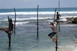 Several individuals are practicing stilt fishing in the ocean, sitting on wooden poles anchored in shallow waters. Waves gently crash in the background, and there is a sense of calm and concentration among the fishermen.
